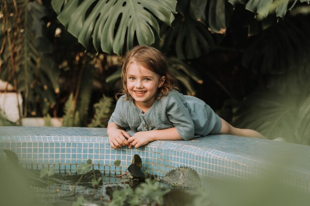 Маленькая блондинка лежит на берегу бассейна с водными растениями в оранжерее сада