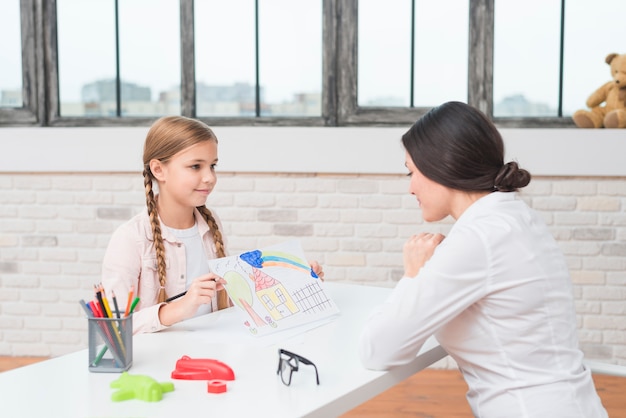 A little blonde girl showing drawn house on paper to her female psychologist