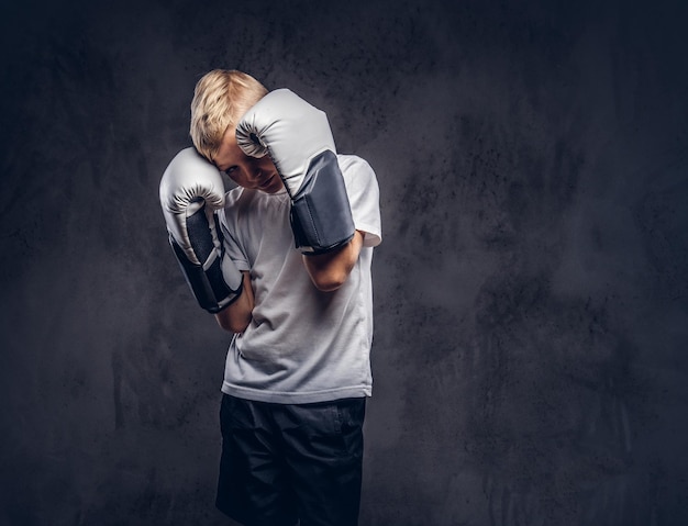 Little boxer with blonde hair dressed in a white t-shirt wearing boxing gloves, workout in a studio. Isolated on a dark textured background.