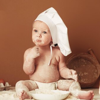 茶色​の​背景​に​小麦粉​で​遊んで​テーブル​に​座っている​シェフ​の​帽子​の​小さな​男の子​の​パン屋