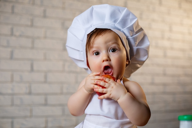 Little boy dressed as a cook in the kitchen. handsome child dressed in an apron eating an apple