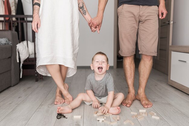 Little boy having fun with wooden blocks with his parents standing behind him holding each other's hand