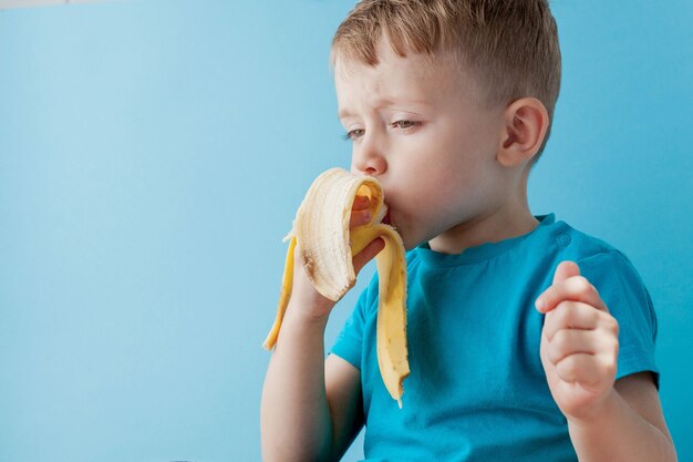 어린 소년 들고 파란색 벽, 음식, 다이어트 및 건강 한 먹는 개념에 바나나를 먹고.