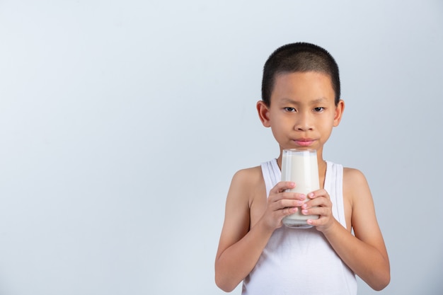 Little boy is drinking glass of milk on white wall.