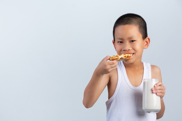Little boy is eating waffles with glass of milk on white wall.
