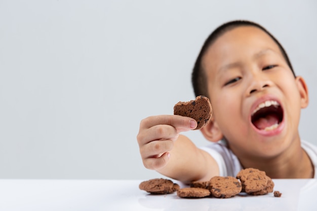 小さな男の子は白い壁のテーブルで食べたいクッキーを持っています。