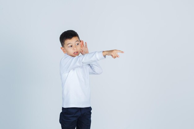 Маленький мальчик держит руку за ухом, указывая в сторону в белой рубашке, штанах и с любопытством смотрит. передний план.