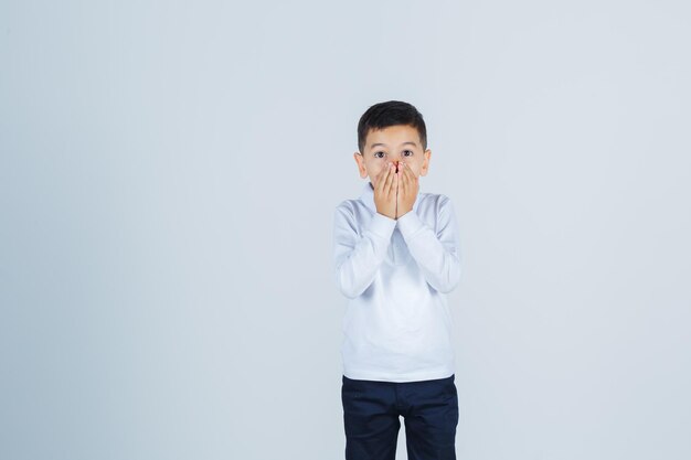 Little boy keeping hands on mouth in white shirt, pants and looking excited , front view.