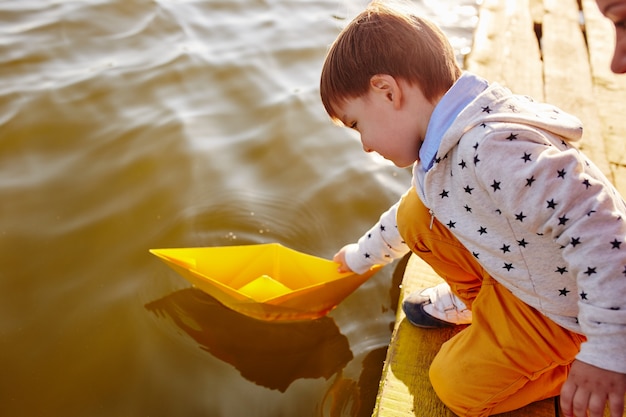 Little boy playing with toy paper ship by the lake