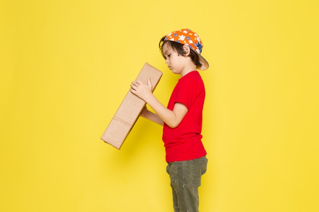 Free photo little boy in red t-shirt and colorful cap holding box on the yellow wall