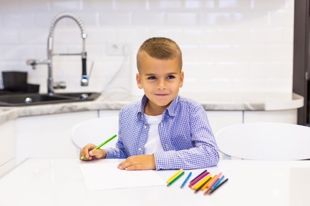 Little boy sits at a table in a bright kitchen and draws with pencils