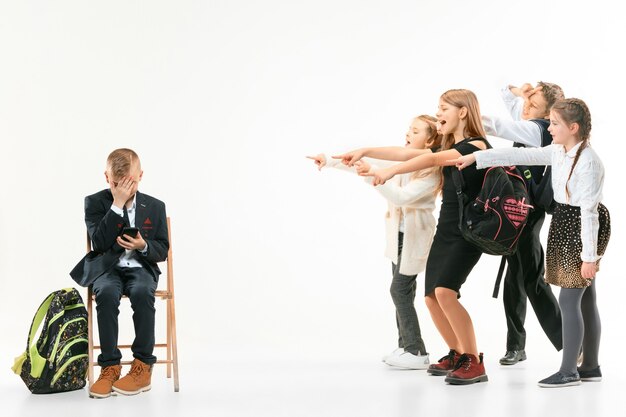 Little boy sitting alone on chair and suffering an act of bullying while children mocking. Sad young schoolboy sitting on  against white wall.