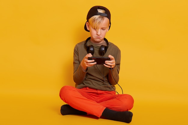 ズボン、帽子、ジャンパーを身に着けている手でスマートフォンを床に座っている男の子、金髪の男性の子供は彼のお気に入りのオンラインゲームをプレイして集中して見えます。子供の頃のコンセプトです。