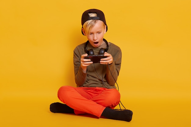 Little boy sitting with smartphone in, guy wearing casually with headphones around neck, posing withopened mouth and looks excited, child with crossed legs, holding mobile phone in hands.