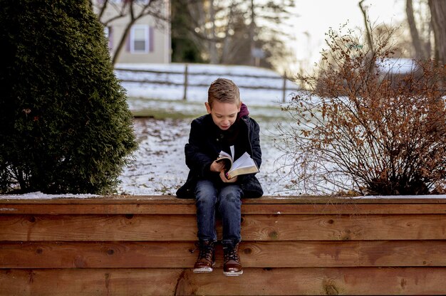 木の板の上に座って、雪に覆われた庭で聖書を読んでいる少年