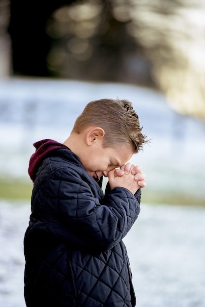 Little boy standing in a park and praying under sunlight with a blurry background