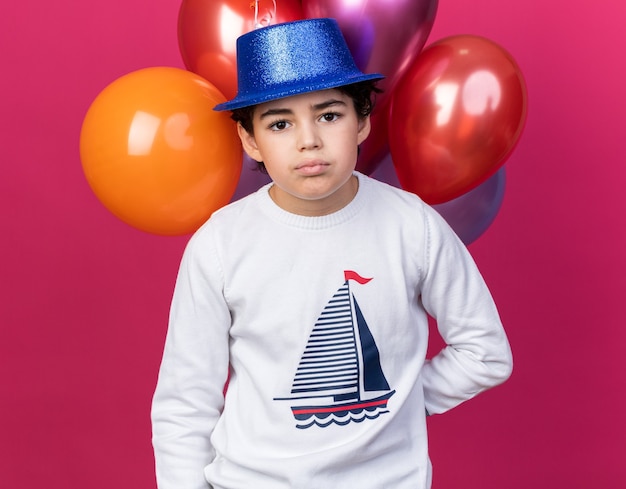 little boy wearing blue party hat standing in front balloons isolated on pink wall