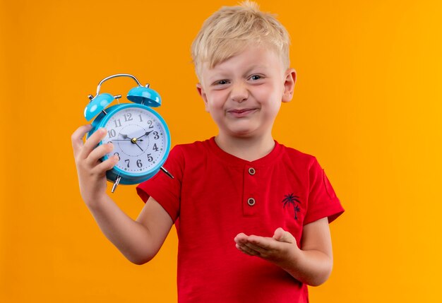 A little boy with blonde hair and blue eyes wearing red t-shirt showing blue alarm clock while looking on a yellow wall