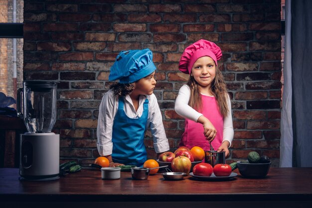 갈색 곱슬머리에 파란색 요리사 유니폼을 입은 어린 소년과 분홍색 요리사 유니폼을 입은 아름다운 소녀가 벽돌 벽에 기대어 부엌에서 함께 요리를 하고 있습니다. 귀여운 꼬마 요리사 커플.