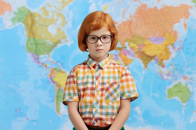 市松模様のシャツと眼鏡を身に着けている地図に立って、学校に行く生姜髪の小さな男の子。賢い生徒が学校の地理キャビネットに立ってレッスンを受ける