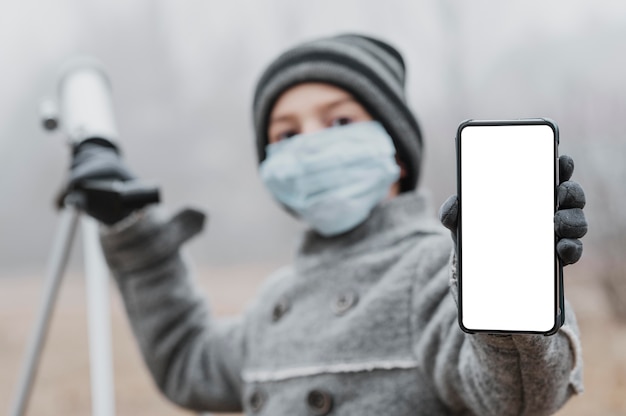 Free photo little boy with medical mask using a telescope and holding a blank smartphone