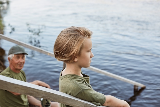 Ragazzino con vista misteriosa guardando in lontananza, ragazzo con il nonno in posa sulle scale in legno, famiglia trascorrere del tempo insieme all'aria aperta, godendo della splendida natura.