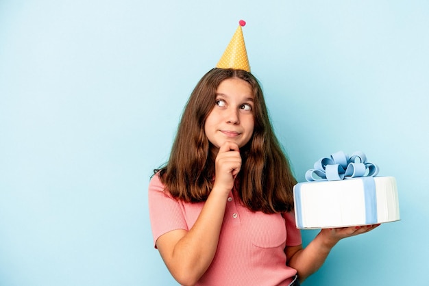 Little caucasian girl celebrating her birthday holding a cake isolated on blue background looking sideways with doubtful and skeptical expression.