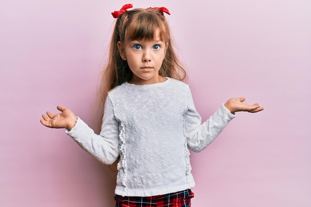 カジュアルな服を着た小さな白人の女の子の子供は、無知で両手を広げて混乱し、考えもなく、疑わしい顔をしています。