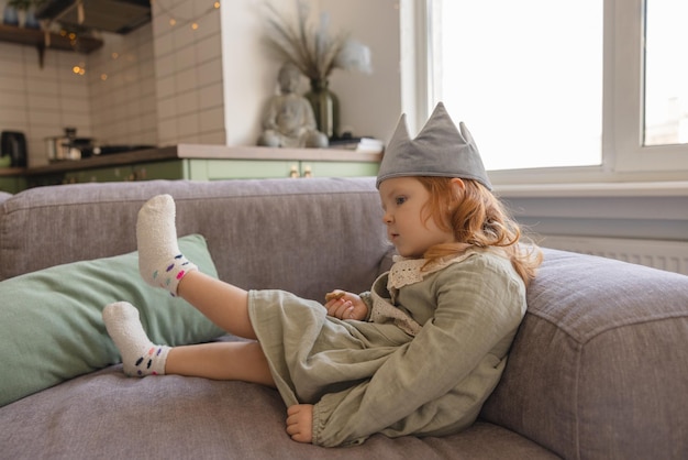 小さな白人の赤毛の少女は、部屋のソファに座っている間、ドレスソックスとおもちゃの王冠を身に着けています子供のライフスタイルの概念