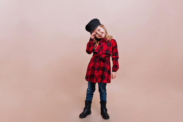 매력적인 소녀는 체크 무늬 셔츠, 청바지와 세련된 모자를 입고 진정한 행복한 감정을 가진 격리 된 벽에 포즈를 취합니다.
