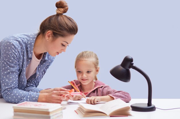 little charming girl sits at table, has difficult homework task, her mother trying to help daughter and explains mathematics rules, uses reading lamp for good vision. Education concept.