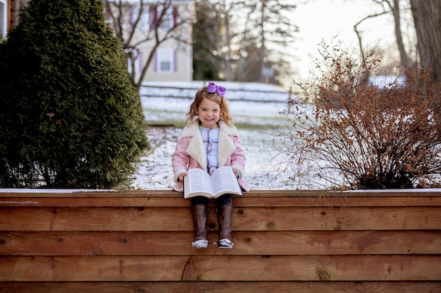 Free photo little cheerful smiling girl sitting on the wooden porch and holding a book