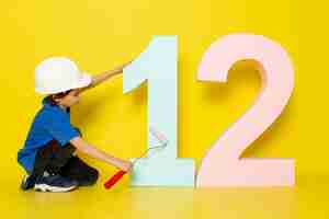 Free photo little child boy in blue t-shirt and white helmet touching number figure on yellow wall