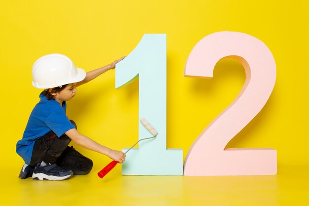 little child boy in blue t-shirt and white helmet touching number figure on yellow wall