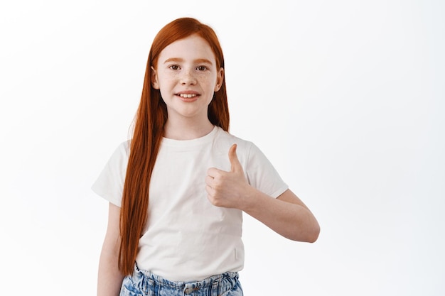 Little child girl with long ginger hair, redhead kid shows thumbs up and smile, nod in approval, praise good thing, like something, make compliment, white background
