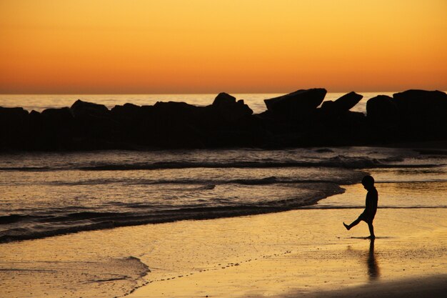 Little child plays on the ocean shore standing before the waves in lights of sunset