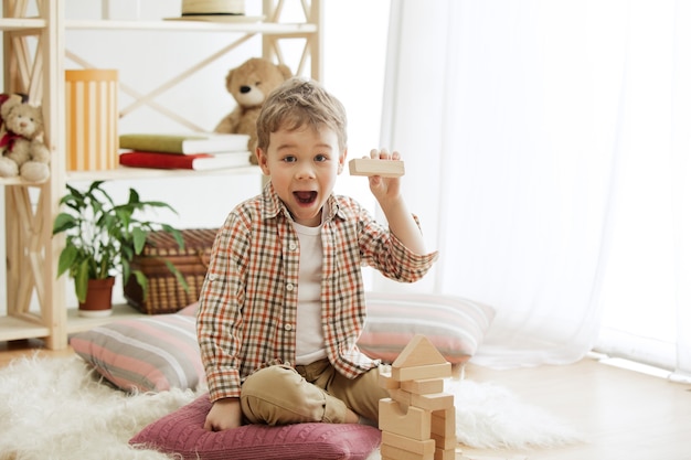 Little child sitting on the floor. Pretty smiling surprised boy palying with wooden cubes at home.