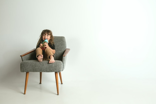 Little child sitting and playing in armchair isolated on white studio wall