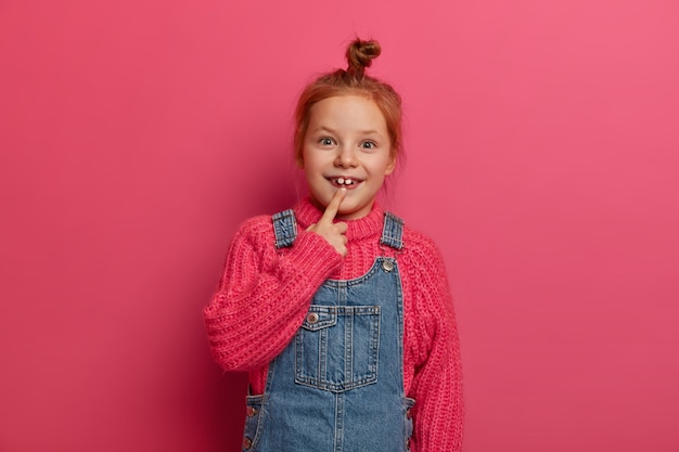 Little child with ginger hair knot points at two adult teeth, has glad expression, wears knitted sweater and denim sarafan, has positive mood, poses against rosy wall. Childhood concept