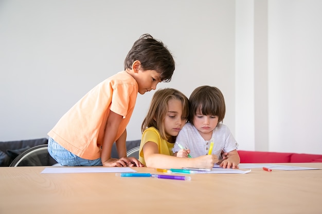 Little children painting with markers in living room. Cute blonde girl holding brother. Lovely kids sitting at table, drawing on paper and playing at home. Childhood, creativity and weekend concept