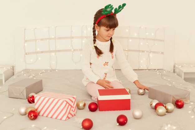 Little concentrated girl with pigtails wearing white sweater and party green deer horns playing on bed with Christmas balls and garland, playing alone, celebrated New Year.