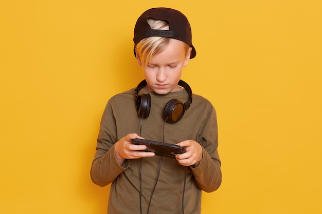Маленький сконцентрированный парень в повседневной рубашке и кепке, играющий в онлайн-видео игры с помощью мобильного телефона, позирующий в наушниках