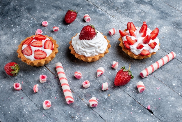 Piccole torte cremose con fragole affettate e fresche insieme a caramelle stick sulla scrivania grigia, colore dolce torta di frutta