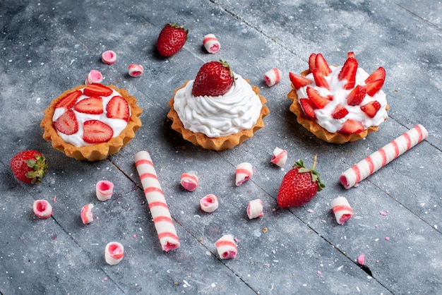 little creamy cakes with sliced and fresh strawberries along with stick candies on grey desk, fruit cake sweet color