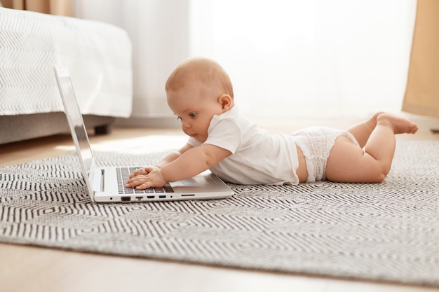 Little curious child studying modern technology while lying on floor on tummy against window, toddler using laptop at home, infant wearing white t shirt.