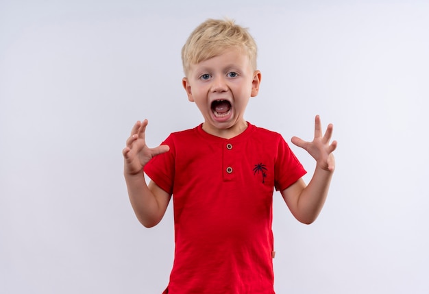 A little cute blonde boy in red t-shirt screaming with his hands up while looking on a white wall