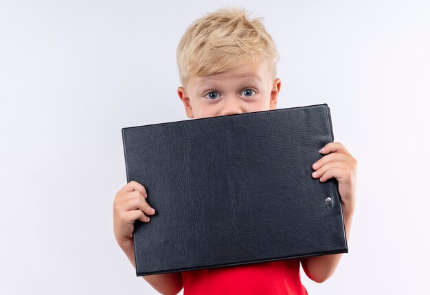 A little cute blonde boy in red t-shirt smiling and holding blank folder while looking on a white wall