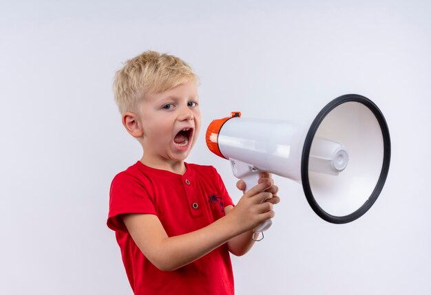 A little cute blonde boy in red t-shirt speaking through megaphone while looking on a white wall