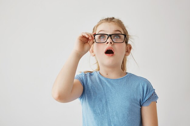 Little cute blonde girl with lovely blue eyes in blue t-shirt and with glasses looks up side opening mouth in surprise seeing a big spider on wall.