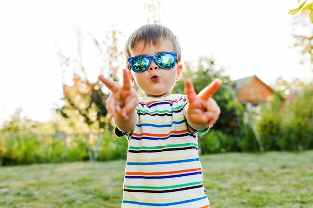Маленький милый мальчик весело и очень серьезно выглядит в своих солнцезащитных очках в саду.